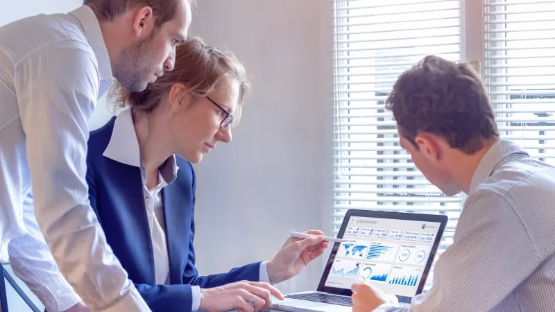 Three business people in office looking at the laptop with dashboard on the screen.