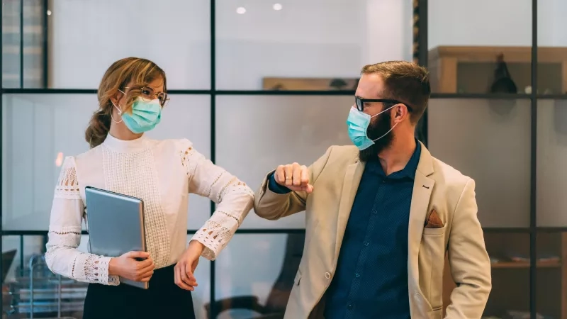 A man and woman wearing masks standing in an office while shaking hands and smiling.
