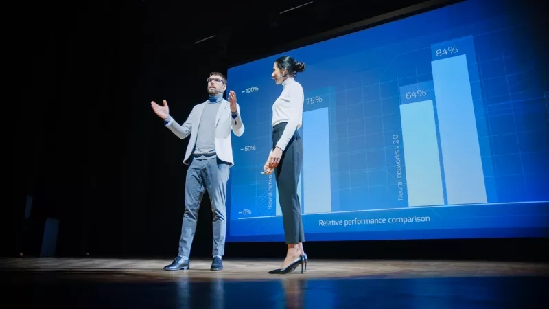 A man and a woman standing on stage in front of a large screen with a graph.