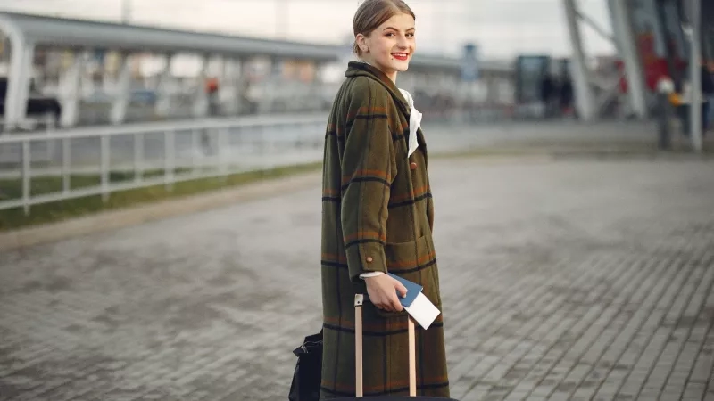 A woman at the airport holding her luggage and smiling for the camera in front of her.