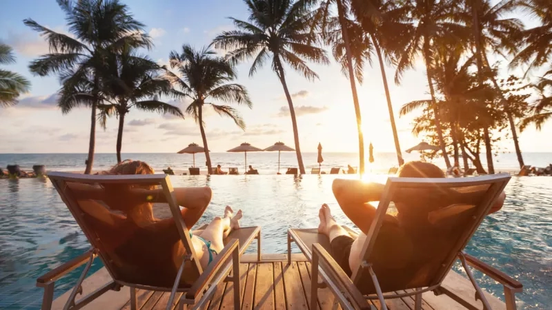 Two tourists sitting by pool at tropical beach with palm trees and sun rays in the sky.