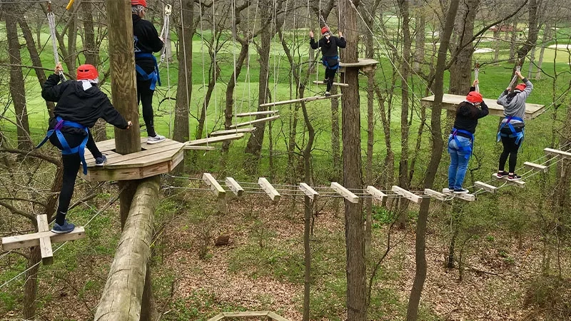 Group of five people navigating an obstacle course with wooden planks and rope bridges in the woods.
