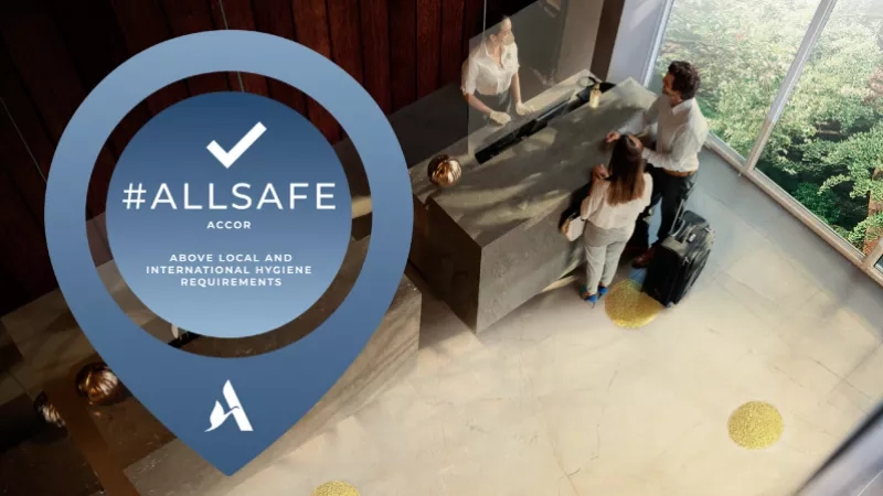 A hotel lobby with #AllSafe branding in the foreground and guests in the background.