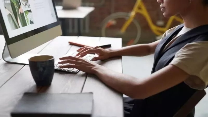 A woman is working at a desk with a computer, a cup, and a book.