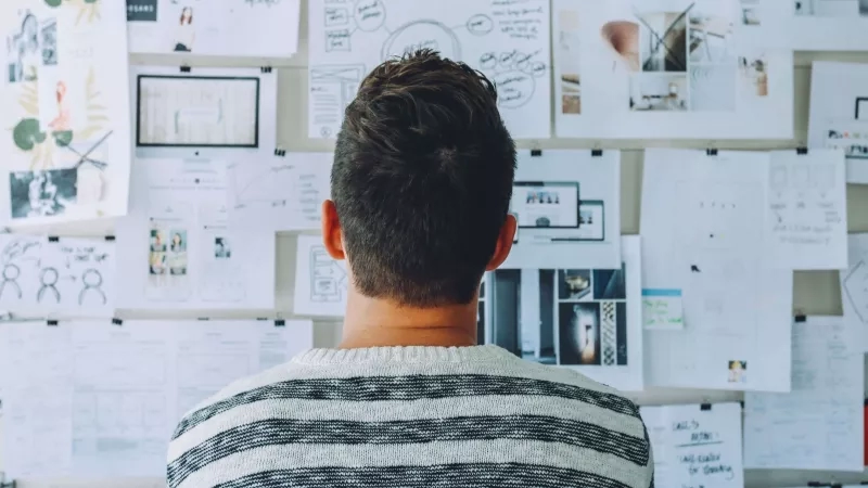 A man is looking at the wall with papers attached to it with words like "object," "text," and "architecture."