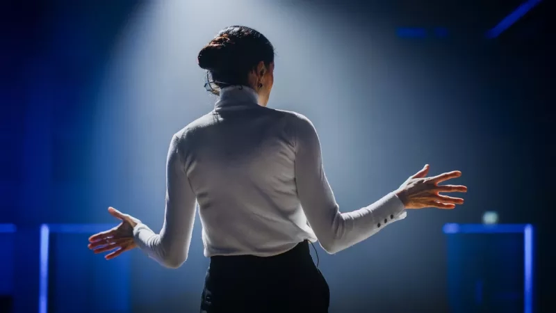 Woman presenting on stage speaking to audience at conference, back view of woman with hands outstretched.