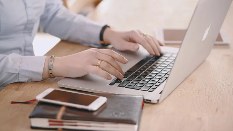 A woman typing on a laptop while a cellphone and a book are beside her.