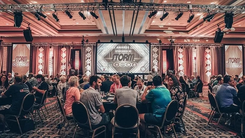 People sit at tables in a conference room with a large screen showing "Storm" text and stage lights.