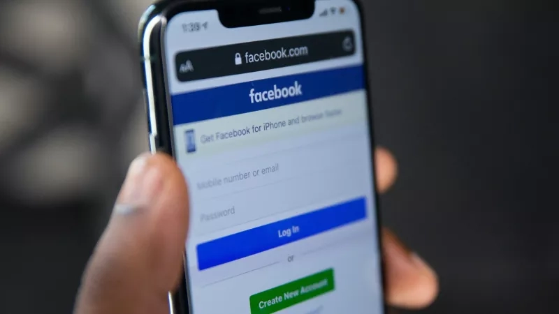 A hand holds a smartphone displaying the Facebook login screen with options to log in or create a new account.