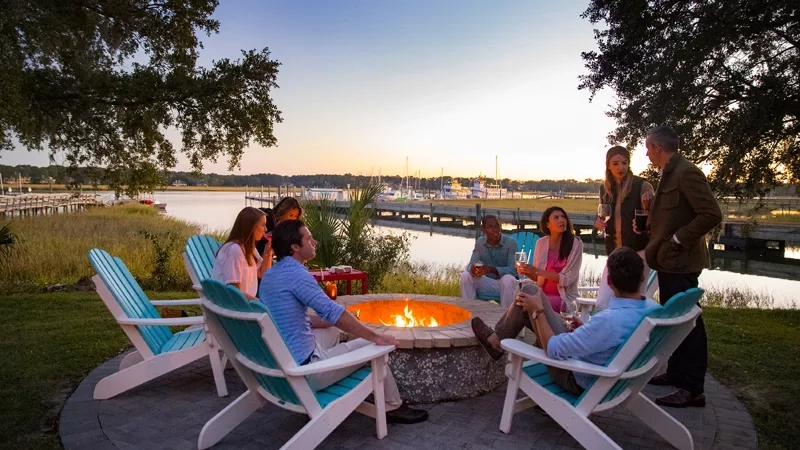 Group of eight adults sit around a fire pit with drinks and a view of the water.