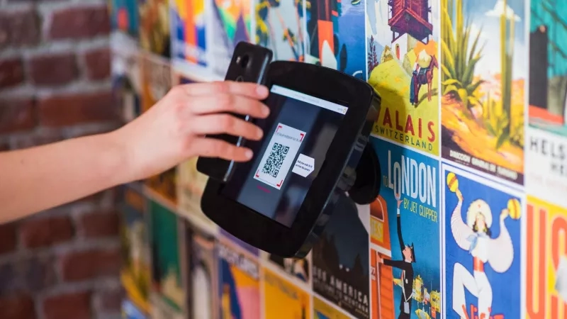 A hand scans a QR code on a Proxyclick Visitor Management System device against a wall of posters.