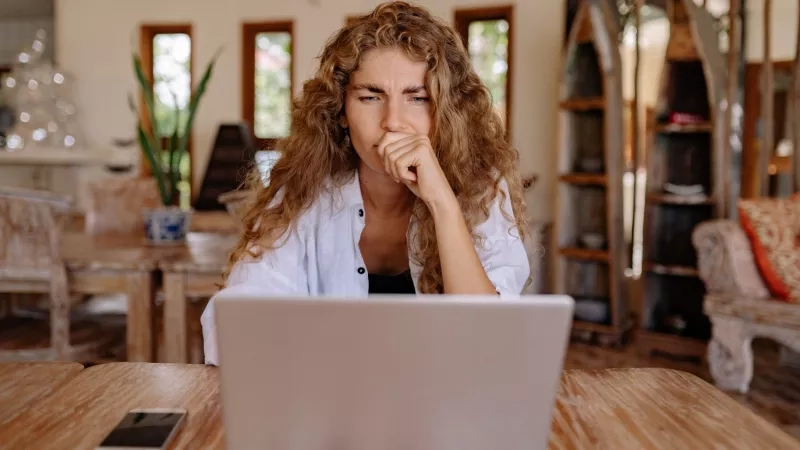 Woman at desk with laptop and phone, hand on face, in a homey room with wooden furniture.