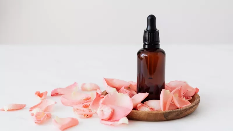 A bottle of rose oil with pink petals on a wooden tray on a white surface.