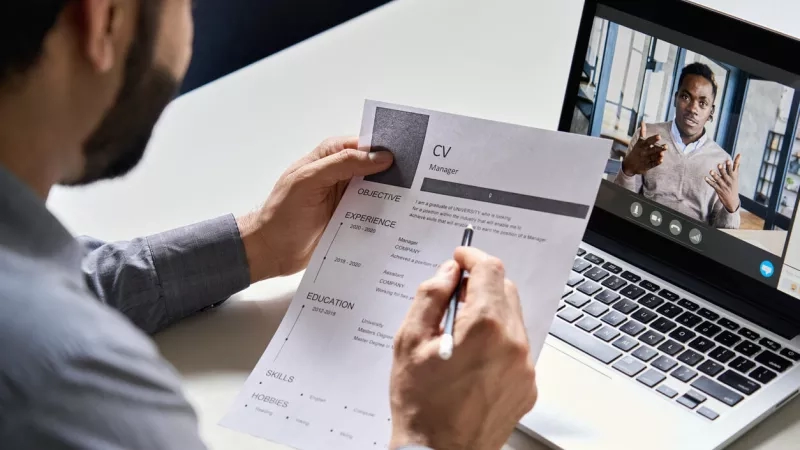 Hiring manager reading resume during video interview with candidate on laptop screen.
