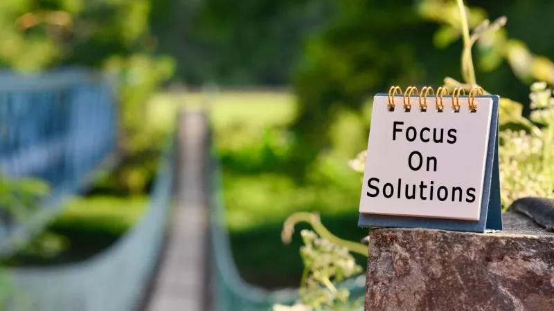 Notepad with inspirational quote "Focus on Solutions" written on it in front of blurred park landscape.