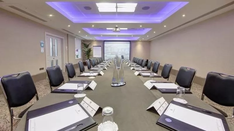Meeting room with table, chairs, books, pens, glass, paper, projector, and ceiling lights in Hilton London Paddington.