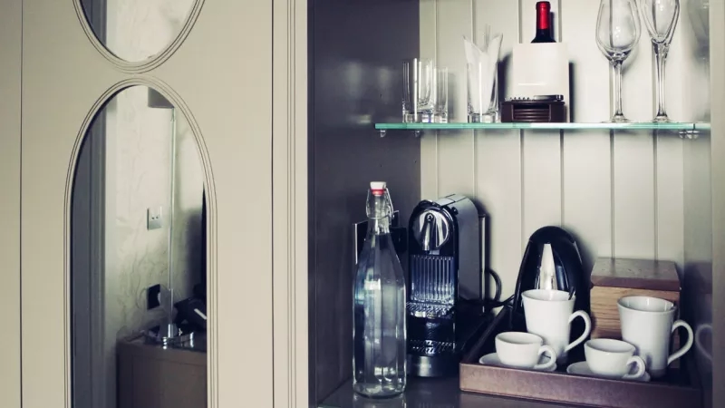 Mini bar with a coffee machine, red wine, and glassware inside a hotel room.