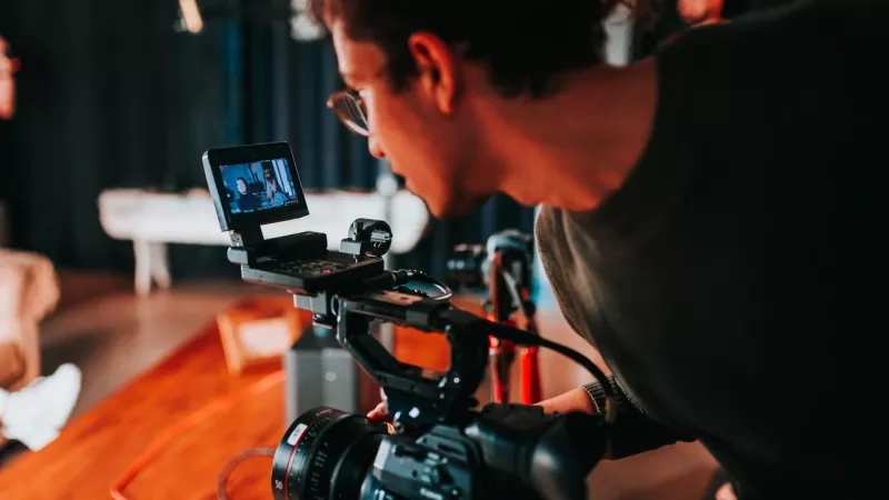 Person with glasses and dark shirt operating camera with small screen and cord on wooden table.
