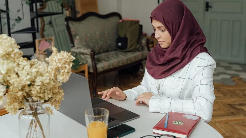 A woman wearing hijab is working on a laptop on a table with glasses, book, and glass of orange juice.