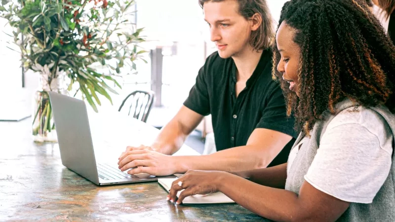 Two people work together on a laptop in a bright, inviting room with flowers and a wooden table.