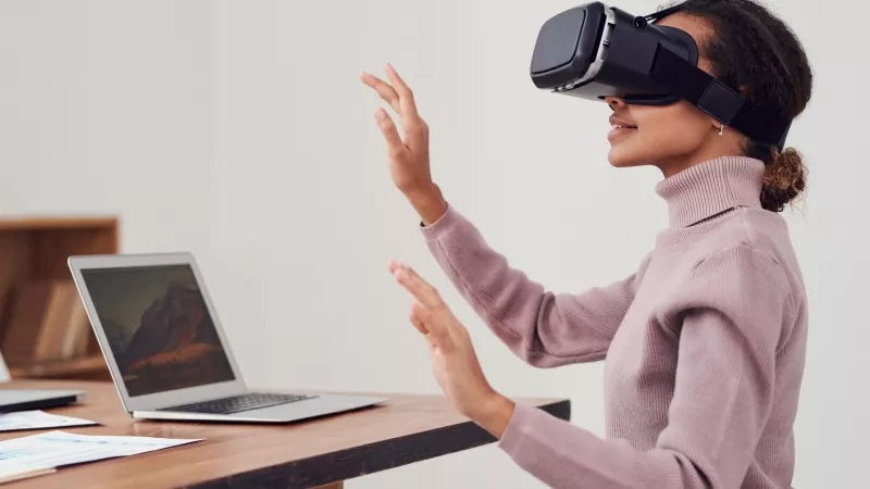 A woman wearing a VR headset is sitting at a desk with a laptop and papers.