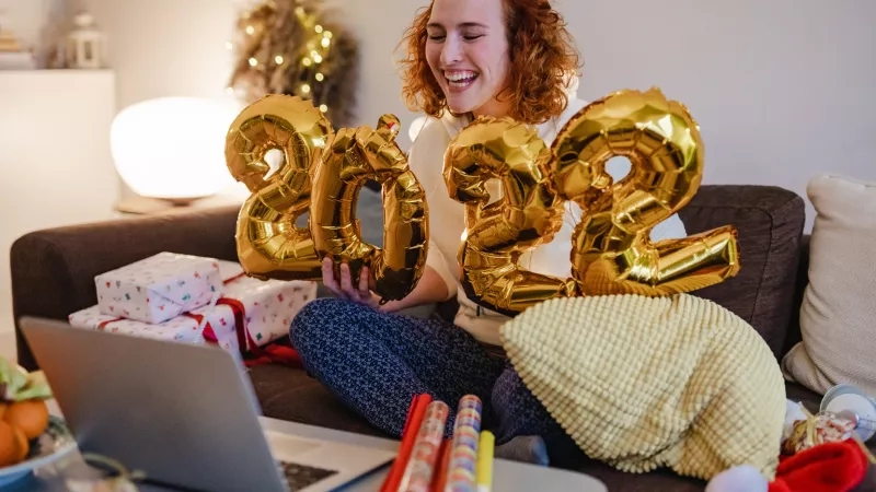 Woman with number balloons celebrating new year on couch with laptop and gifts, festive decor in background.