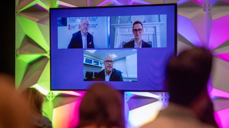 Three men appear on a screen as part of a video conference, with two people watching.