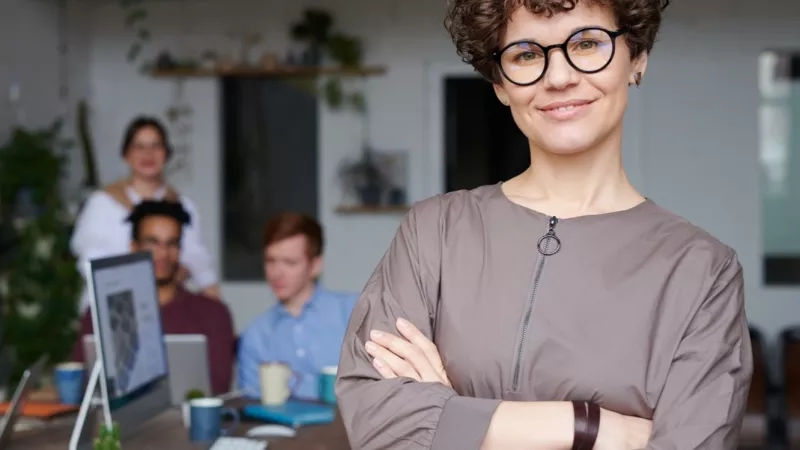 A woman stands smiling in an office with coworkers in the background working at desks.