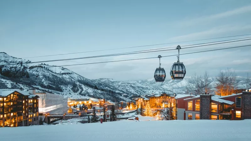 Gondolas in the sky, buildings, and mountains in the distance in the snow.