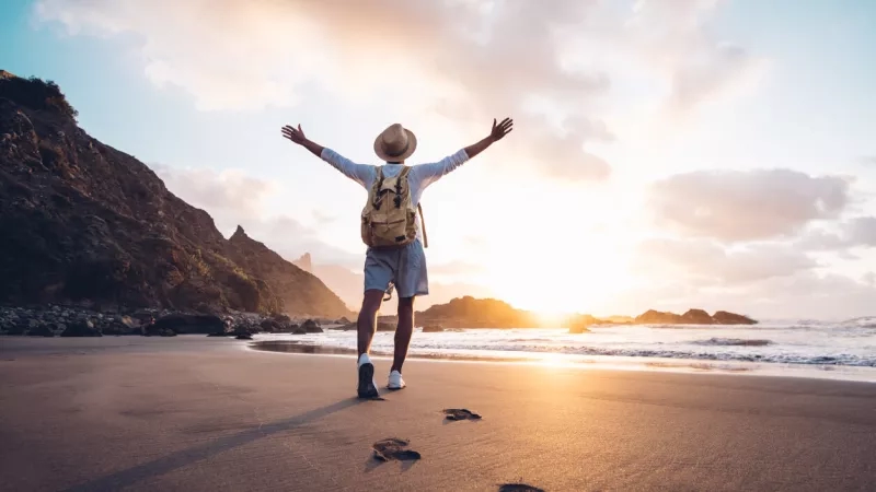 A man with a backpack is walking on the beach at sunset with his arms spread out wide.