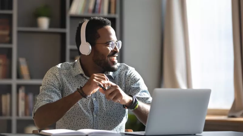 Man sitting at computer smiling with headphones listening to podcast.