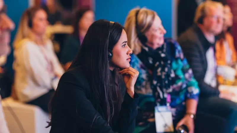 A group of people sit at a conference, one woman in black leans forward with her hand on her chin.