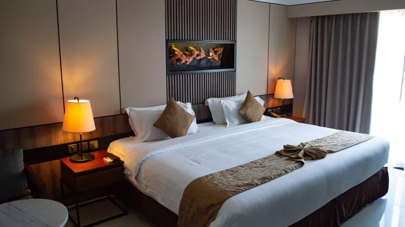 Hotel bedroom interior with large bed, artwork, curtains, lamps, and bedside tables with drawers.