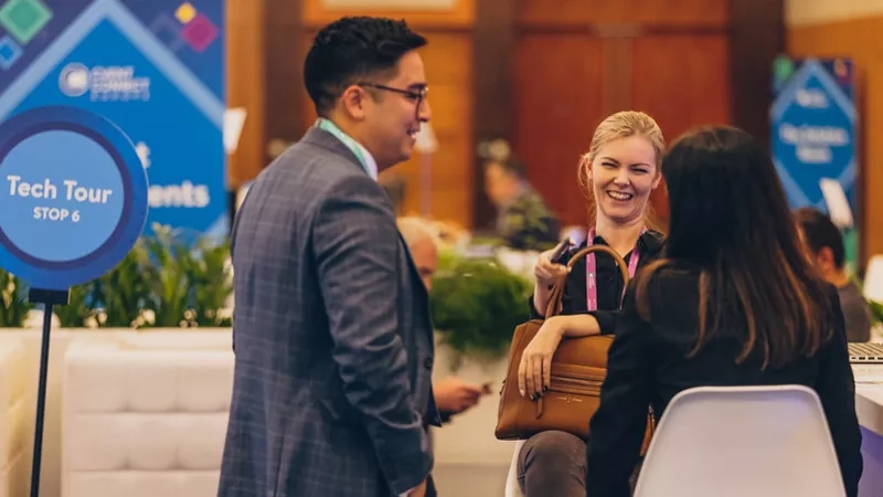 Two people are smiling and talking at a conference with a sign saying Tech Tour Stop 6.