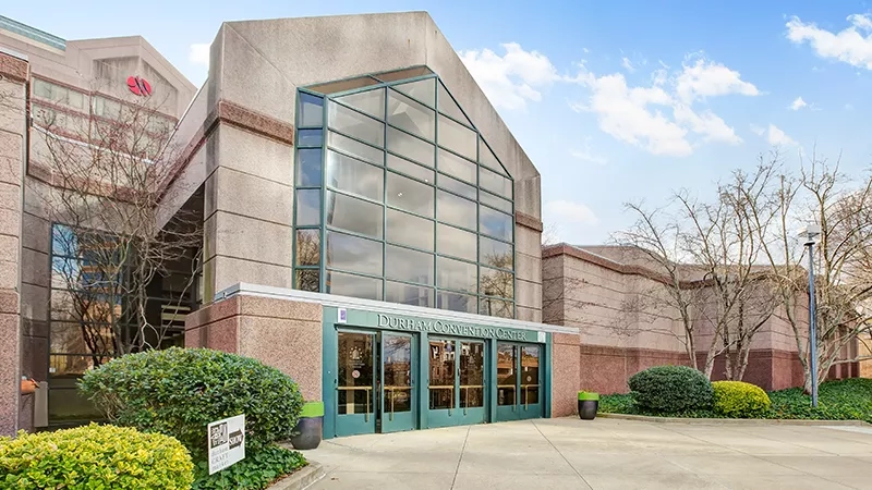 Exterior of Durham Convention Center with glass doors and windows, greenery, and a sign in front.
