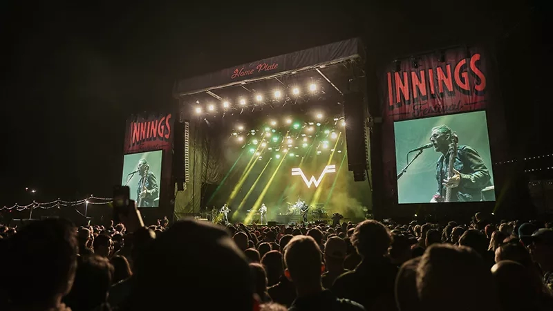 A large crowd at a music festival with large screens showing the band on stage.