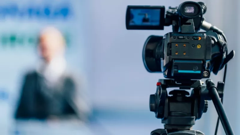 Close up of a video camera on a tripod filming a speaker in a conference room.