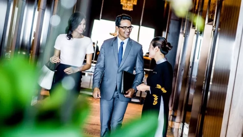Businessman and two women walk in hotel corridor with modern architecture and green plants.