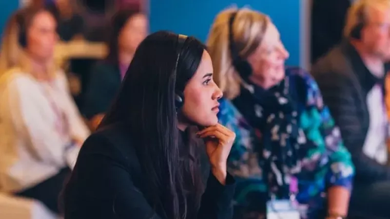 A woman with headphones and her hand on her chin is sitting in a row of people.