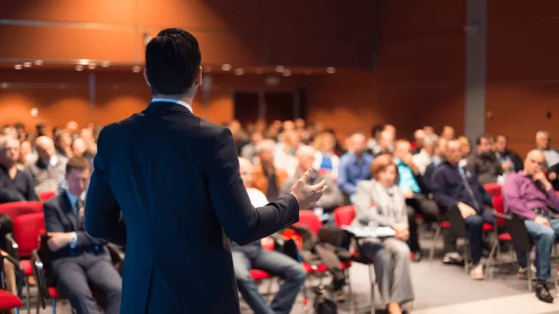 A man giving a presentation in front of a crowd of people in a room with orange walls.