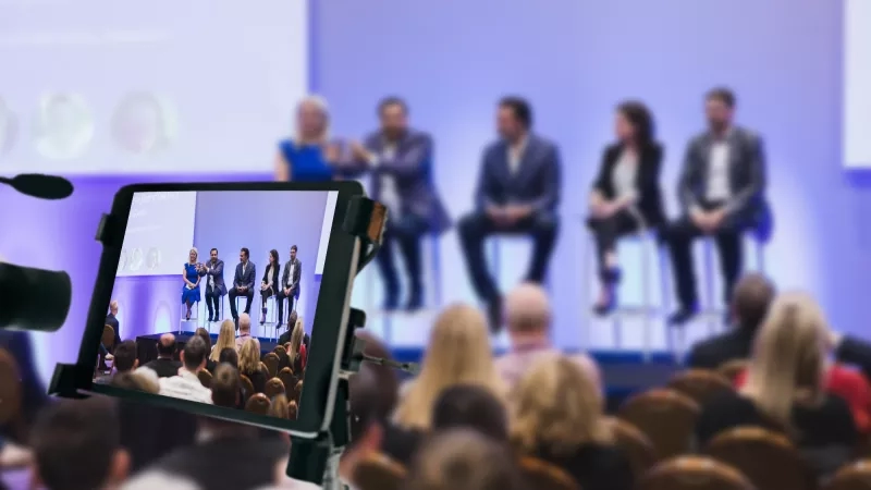 Camera recording a panel of speakers at a conference in front of an audience in a conference hall.