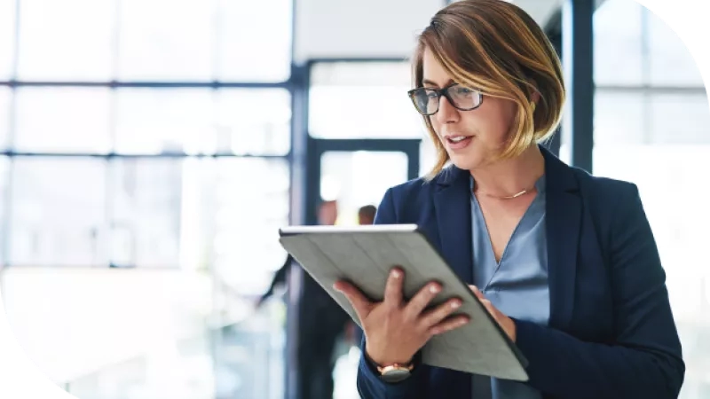 A woman stands in an office, holding a tablet and looking at it while wearing a business suit.