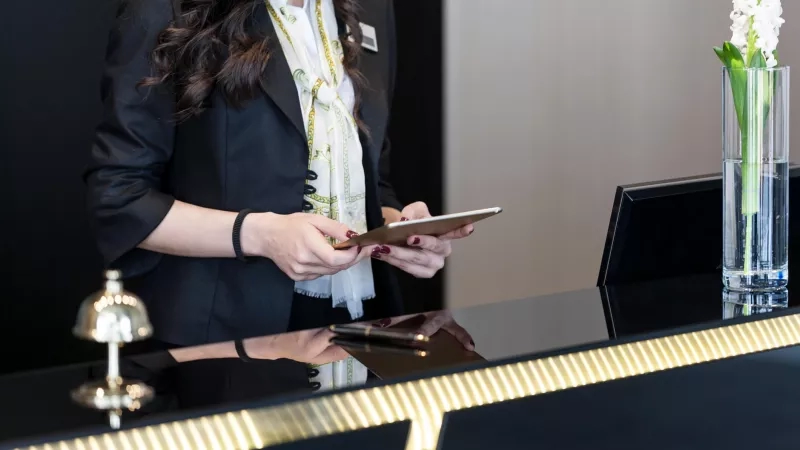 Hotel operations manager reviewing data on tablet at front desk in hotel lobby.
