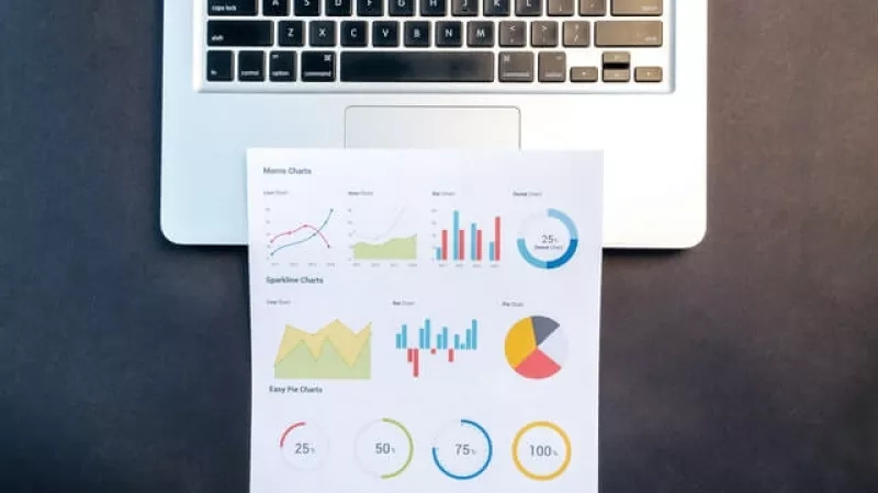 Laptop keyboard with paper with colorful charts on top of it, in front of a gray background.