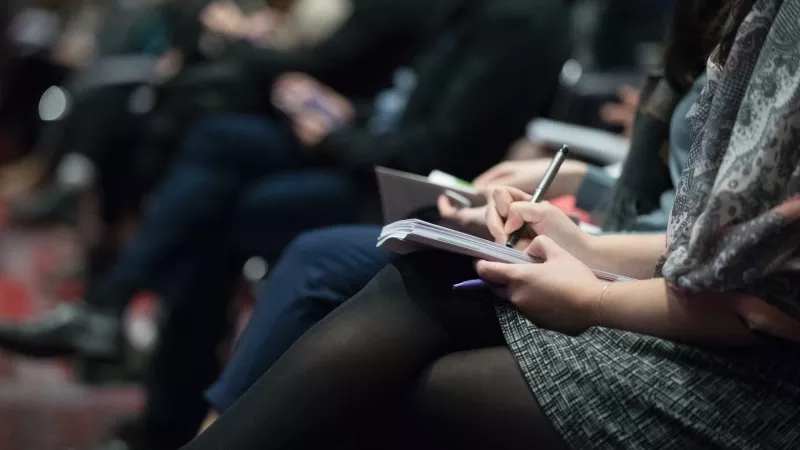 A woman holding a notebook and pen while sitting next to a man in a lecture hall.