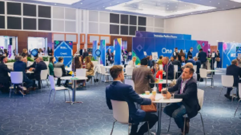 Two business people talking at a conference while others are sitting at the tables.