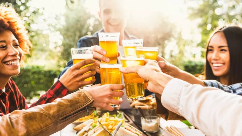 Group of people toasting with beers at brewery event during summer with sun flare in background.