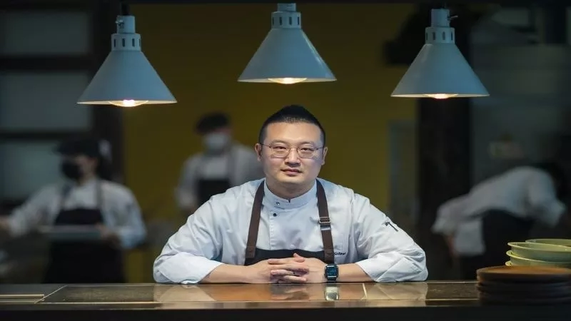 Chef standing in front of a counter in the kitchen with other chefs in the background.