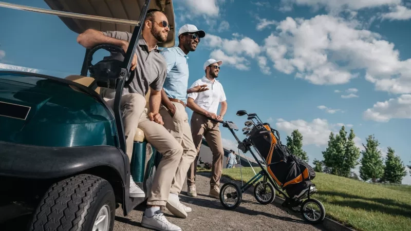 Three men chatting during a social event at a golf club, with golf carts and clubs nearby.
