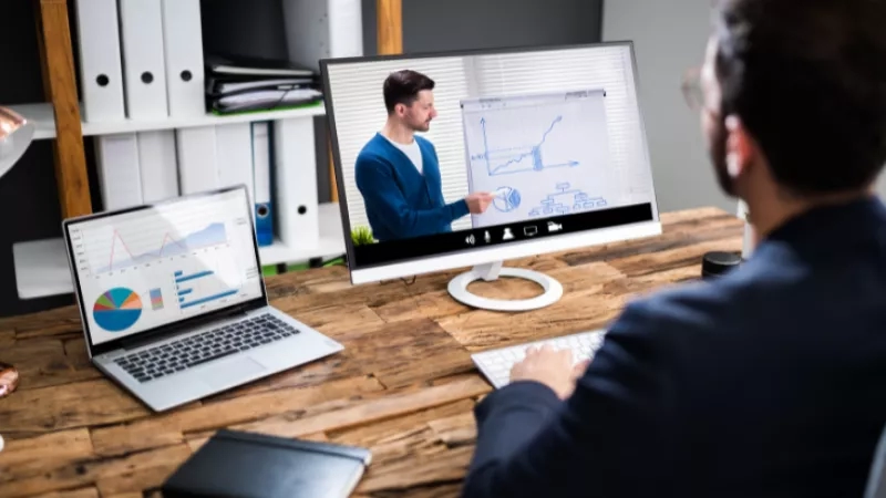 Man in business attire, using a laptop and computer monitor on a wooden desk for a video call.